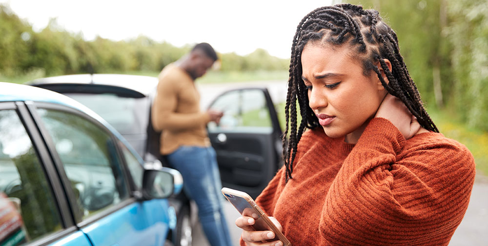 A woman holding her neck and on her cell phone with a man and two cars that crashed in the background